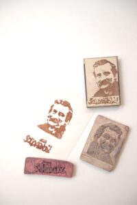 Zbiór pieczątek ze stanu wojennego z wizerunkiem Lecha Wałęsy. Materiał: gumka myszka, papier. Data powstania: lata: tysiąc dziewięćset osiemdziesiąt dwa do tysiąc dziewięćset osiemdziesiąt sześć. Wymiary: trzy i sześć dziesiątych centymetra na pięć i trzy dziesiąte centymetra. Kolorowa fotografia pieczątek wykonanych z gumki do mazania oraz ich odbitek na niewielkim, prostokątnym kawałku papieru i pudełku od zapałek. Pieczątki są dwie: jedna w kształcie prostokąta z precyzyjnie wyciętym negatywem napisu: Solidarność wykonanego charakterystyczną czcionką nazwaną później „solidarycą”. Litery są nierówne, mają nieregularne kształt, niektóre łączą się ze sobą. Z litery N wystaje maszt, na którego szczycie znajduje się powiewająca biało-czerwona chorągiew. Druga pieczątka z wyciętym negatywem portretu Lecha Wałęsy. Na pieczątce kojarzone z Lechem Wałęsą w latach osiemdziesiątych: sumiasty wąs i i krzaczaste brwi. Obie pieczątki odbite są razem – zarówno na kartce papieru, jak i na pudełku zapałek. Portret Lecha Wałęsy znajduje się powyżej napisu: „Solidarność”.