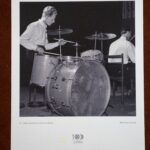 Fotografia, Koncert Dom Kultury Zabrze. Autor: Władysław Dziczyński. Materiał: papier fotograficzny. Miejsce wykonania: Dom Kultury Zabrze, rok tysiąc dziewięćset pięćdziesiąty szósty. Wymiary: dwadzieścia pięć na dwadzieścia centymetrów. Czarno-biała fotografia w białym passe-partout. Na fotografii, po lewej stronie: Adam Jędrzejowski, siedzi przy instrumentach perkusyjnych, w dłoniach trzyma pałeczki. Ma na sobie białą koszulę z długimi rękawami i czarne spodnie. Po prawej stronie, na drugim planie, odwrócony plecami przy fortepianie siedzi Tadeusz Erol Kosiński. Pod fotografią na passe-partout napisy. Z lewej: Adam Jędrzejowski – perkusista jazzowy, po prawej: Władysław Dziurzycki. Pod spodem logo stulecia Zabrza.