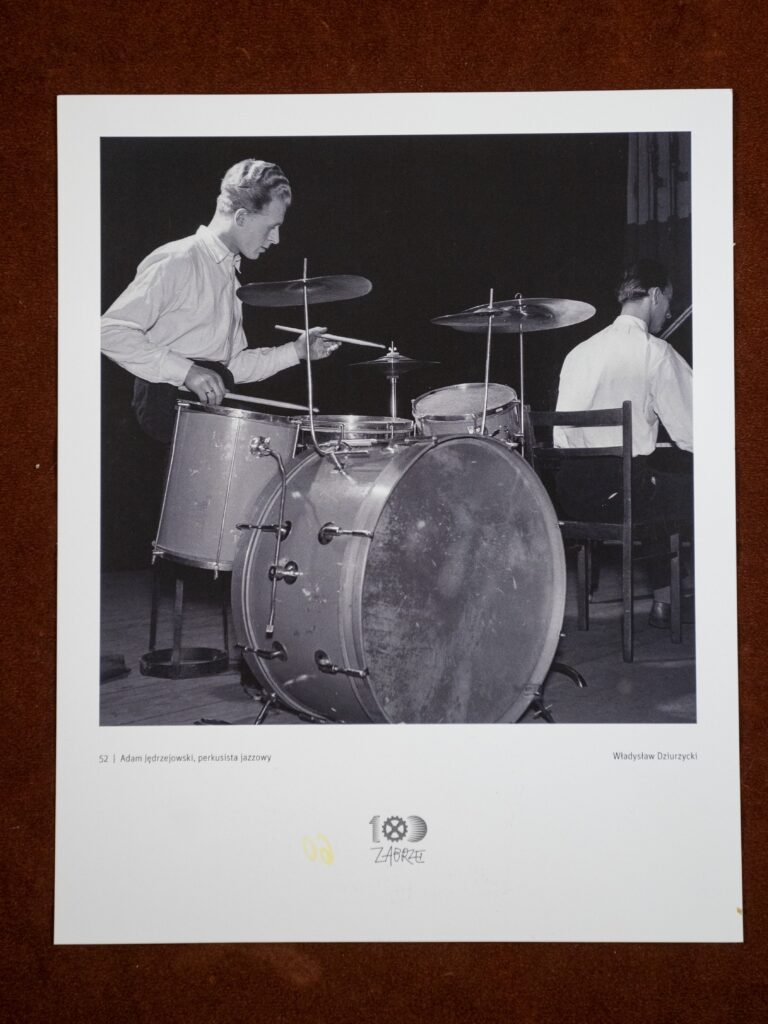 Fotografia, Koncert Dom Kultury Zabrze. Autor: Władysław Dziczyński. Materiał: papier fotograficzny. Miejsce wykonania: Dom Kultury Zabrze, rok tysiąc dziewięćset pięćdziesiąty szósty. Wymiary: dwadzieścia pięć na dwadzieścia centymetrów. Czarno-biała fotografia w białym passe-partout. Na fotografii, po lewej stronie: Adam Jędrzejowski, siedzi przy instrumentach perkusyjnych, w dłoniach trzyma pałeczki. Ma na sobie białą koszulę z długimi rękawami i czarne spodnie. Po prawej stronie, na drugim planie, odwrócony plecami przy fortepianie siedzi Tadeusz Erol Kosiński. Pod fotografią na passe-partout napisy. Z lewej: Adam Jędrzejowski – perkusista jazzowy, po prawej: Władysław Dziurzycki. Pod spodem logo stulecia Zabrza.