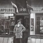 Fotografia: Adam Jędrzejowski przed swoim biurem podróży w Sztokholmie, autor nieznany. Materiał: papier fotograficzny. Sztokholm, tysiąc dziewięćset siedemdziesiąt dwa. Wymiary: dwadzieścia cztery na osiemnaście centymetrów. Ulica przed witryną Biura Podróży. W drzwiach stoi mężczyzna w grubej kurtce zapinanej na kołki, z kołnierza rozchylonej kurtki wystaje jasny, wełniany golf. Ma głowie mężcyzny czapka. Prawą rękę trzyma w kieszeni kurtki. Lewą wkłada lub wyciąga. Patrzy w lewą stronę. Za jego plecami witryna biura podróży. W oknach figurki drewnianych świątków, ikona, plakat z Mikołajem Kopernikiem. Napis u góry: Skanpolbus.