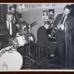 Fotografia z koncertu. Twórca nieznany. Materiał: papier fotograficzny. Miejsce wykonania: Newcastle [Njukastl] rok tysiąc dziewięćset sześćdziesiąt cztery. Wymiary: dwadzieścia dwa na szesnaście centymetrów. Czarnobiałe zdjęcie z koncertu. Na zdjęciu od lewej przy perkusji i bębnach Adam Jędrzejowski. Za jego plecami mężczyzna z kontrabasem. Na prawo od nich, lekko z tyłu przy pianinie Adam Makowicz, najbardziej po prawej na saksofonie gra Zbigniew Namysłowski. Z tyłu na ścianie, za Adamem Makowiczem i fortepianem przyczepione czarno-białe fotografie muzyków.