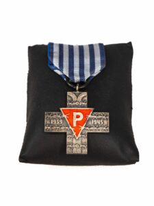 Krzyż Oświęcimski. Materiał: metal. Polska – rok tysiąc dziewięćset osiemdziesiąt pięć. Wymiary czterdzieści dwa na czterdzieści dwa milimetry. Metalowy równoramienny krzyż. Patynowany. W jego centralnej części trójkątny metalowy element – tarcza, koloru czerwonego, jasno obramowana. Na tarczy jasna litera P [Pe]. Powierzchnia ramion krzyża z wzorem w kształcie drutów kolczastych, na pionowym ramieniu krzyża zagięte u góry słupy obozowego ogrodzenia. Na poprzecznym ramieniu krzyża liczby – lata: tysiąc dziewięćset trzydzieści dziewięć – tysiąc dziewięćset czterdzieści pięć. Krzyż oświęcimski przyczepiony do wstążki w kształcie trapezu. Wstążka - w biało niebieskie pasy. Krzyż eksponowany na czarnej poduszce. Fotografia przedstawia awers Krzyża Oświęcimskiego.