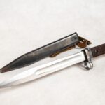 Bagnet. Lata czterdzieste dwudziestego wieku. Brak danych dotyczących długości bagnetu. Bagnet tulejowy z odsłoniętym ostrzem. Obok metalowa pochwa. Na nasadzie metalowej rękojeści wybita liczba: tysiąc dziewięśset pięćdziesiąt osiem. Drewniana nakładka na rękojeść mocowana dwiema śrubami.