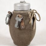 Menażka. Materiał: aluminium. Miejsce wykonania: PRL [ Pe eR eL] , lata czterdzieste dwudziestego wieku. Pojemność jeden i dwie dziesiąte litra. Aluminiowa manierka. Zakręcana aluminiowym wieczkiem. Manierka w sznurowanym pokrowcu z grubej tkaniny. Do pokrowca umocowane napami skórzane pętle z aluminiowymi karabińczykami.