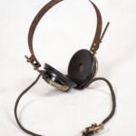 Słuchawki. Wytwórca: Defeton. Materiał: metal. Polska, rok tysiąc dziewięćset dwudziesty dziewiąty. Słuchawki nauszne, otwarte, na metalowym pałąku. Pałąk pokryty cienką warstwą rdzy. Elementy emitujące dźwięk - okrągłe, ich wewnętrzna powierzchnia pokryta tkaniną z niewielkim otworem pośrodku. Od każdej ze słuchawek odchodzą kable, pokryte lekko zetlałą, tkaną izolacją. Końcówki kabli zerwane.