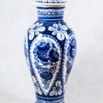 Wicek. Producent: Włocławska Fabryka Fajansu. Materiał: fajans. Wazon w kształcie amfory z fajansowym wieczkiem. Naczynie z okrągłą szyjką nakrytą wypukłym wieczkiem z walcowatym uchwytem. Poniżej szyjki, boki naczynia najpierw uwypuklają się, potem zwężają ku okrągłej podstawie. Fajans jest biały, malowany we wzory kwiatowe w kolorze kobaltowym. Wzory namalowane są na obwodzie szyjki i wokół podstawy. Motywy na bokach naczynia powtarzają się pięciokrotnie. Kwiatowe wzory umieszczono w obramowaniu w kształcie kropli zwężającej się do dołu w kierunku podstawy wazonu.