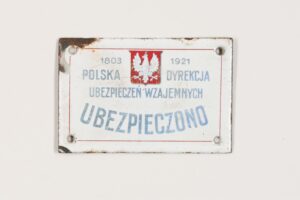 Tabliczka ubezpieczeniowa. Stalowa blacha, emalia piecowa, wykonana techniką wypalania i polerowania w Lesznie w tysiąc dziewięćset dwudziestym pierwszym roku. Wymiary: dziewięć na sześć centymetrów. Tabliczka pokryta białą emalią, z czterema otworami umieszczonymi w pobliżu czterech narożników. Lewy, górny róg uszkodzony: brak emalii, pojawia się rdza. Na tabliczce, w górnej części biały orzeł na czerwonym tle. Herb Polski z białym orłem z lat tysiąc dziewięćset dziewiętnaście – tysiąc dziewięćset dwadzieścia siedem. Napis od góry: tysiąc osiemset trzy – tysiąc dziewięćset dwadzieścia jeden; pod spodem: Polska Dyrekcja; w kolejnym wierszu: Ubezpieczeń Wzajemnych. W dolnej części tabliczki napis umieszczony łukowato: Ubezpieczono.