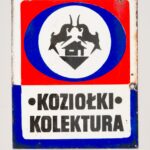 Reklama semaforowa gry liczbowej „Koziołki”. Materiał: blacha stalowa, emalia piecowa. Technika: wypalanie i polerowanie. Leszno, rok tysiąc dziewięćset sześćdziesiąty. Wymiary: dwadzieścia dziewięć i pół na trzydzieści sześć i pół centymetra. Na czerwonym tle w niebieskim obramowaniu sylwetki dwóch, bodących się koziołków, pod nimi sylwetka domku jednorodzinnego. Ta część stanowi około dwóch trzecich tabliczki od góry. W dolnej, jednej trzeciej wysokości tabliczki: białe tło z niebieskim obramowaniem i napisem: Koziołki – kolektura.