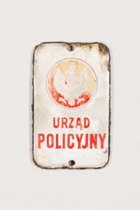 Szyldzik drzwiowy. Projekt rządowy. Emaliernia. Stalowa blacha emaliowana. Wykonano techniką wypalania i polerowania w Lesznie w tysiąc dziewięćset dziewiętnastym roku. Wymiary: sześć centymetrów na dziewięć i osiem dziesiątych centymetra. Tabliczka z emalią lekko uszkodzona na brzegach, szczególnie w górnej części. Na białym tle herb Polski z białym orłem z lat tysiąc dziewięćset dziewiętnaście – tysiąc dziewięćset dwadzieścia siedem, w zamkniętej koronie z wydłużonymi w górę skrzydłami i krótszymi łapami. Orzeł na okrągłej tarczy. Pod herbem napis: Urząd Policyjny.