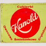 Reklama Kanold Leszno. Stalowa blacha pokryta farbą. Technika nadruku. Leszno, rok tysiąc dziewięćset dwudziesty szósty. Wymiary: dwadzieścia dwa na siedemnaście i pół centymetra. Prostokątna tabliczka z czterema niewielkimi otworami w narożnikach. Obrzeże tabliczki tworzy lekko wklęsłe obramowanie. Większa część tabliczki jest lekko wypukła. Na okrągłym, czerwonym tle, poprowadzony ukosem, ozdobny napis: „Kanold”. Nad kołem ułożony poziomo napis: „Cukierki”. Pod czerwonym kołem od lewej ukosem: „owocowe”, dalej w prawo poziomy napis: „Śmietankowe”, po prawej stronie ukosem: „Grylażowe”.