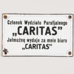 Tabliczka informacyjna Caritas [Karitas]. Materiał: stalowa blacha, emalia piecowa. Technika: wypalanie i polerowanie. Leszno, rok tysiąc dziewięćset trzydziesty. Wymiary: piętnaście na dziewięć centymetrów. Prostokątna tabliczka, na białym tle czarny napis. W czterech narożnikach tabliczki otwory. W okolicach utworów emalia lekko uszkodzona. Napis na tabliczce: „Członek Wydziału Parafjalnego „Caritas”. Jałmużnę wydaje za mnie biuro „Caritas”.
