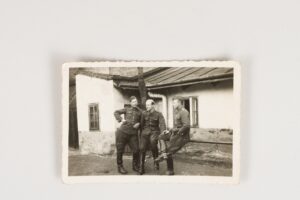 Czarno-biała fotografia. Ułani, dziewiąty pułk strzelców konnych, autor nieznany, Miejsce powstania: Grajewo – koszary, rok tysiąc dziewięćset trzydziesty drugi lub tysiąc dziewięćset trzydziesty trzeci. Wymiary osiemdziesiąt osiem na sześćdziesiąt milimetrów. Trzech mężczyzn w mundurach, furażerkach i wysokich butach. Dwóch przysiadło na koniowiązie, jeden opiera się lewym bokiem. Za nimi drzewo i parterowy dom z oknami i bielonymi ścianami.